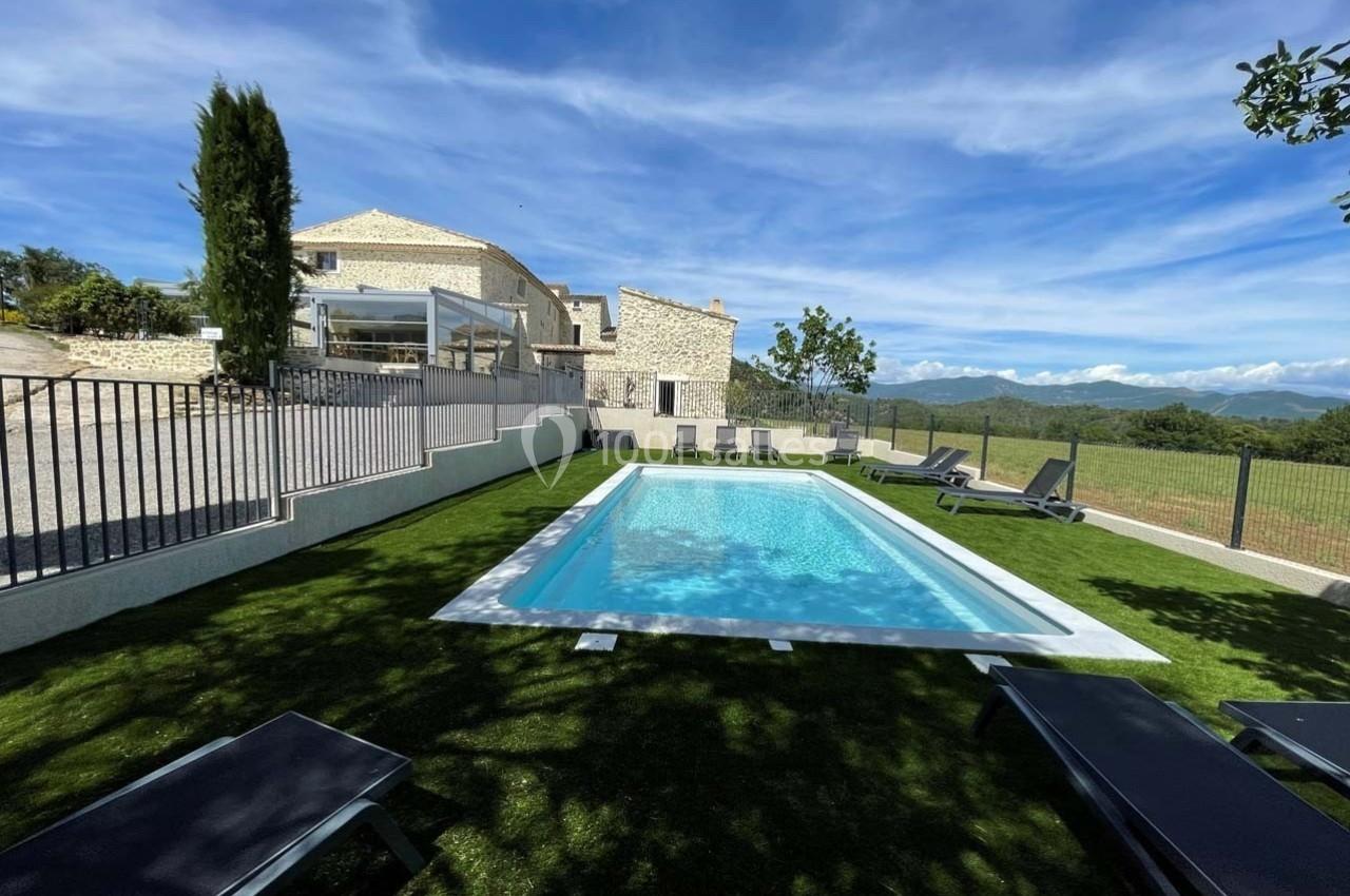 Piscine extérieure rectangulaire entourée de chaises longues, avec vue sur une maison en pierre et un paysage montagneux.