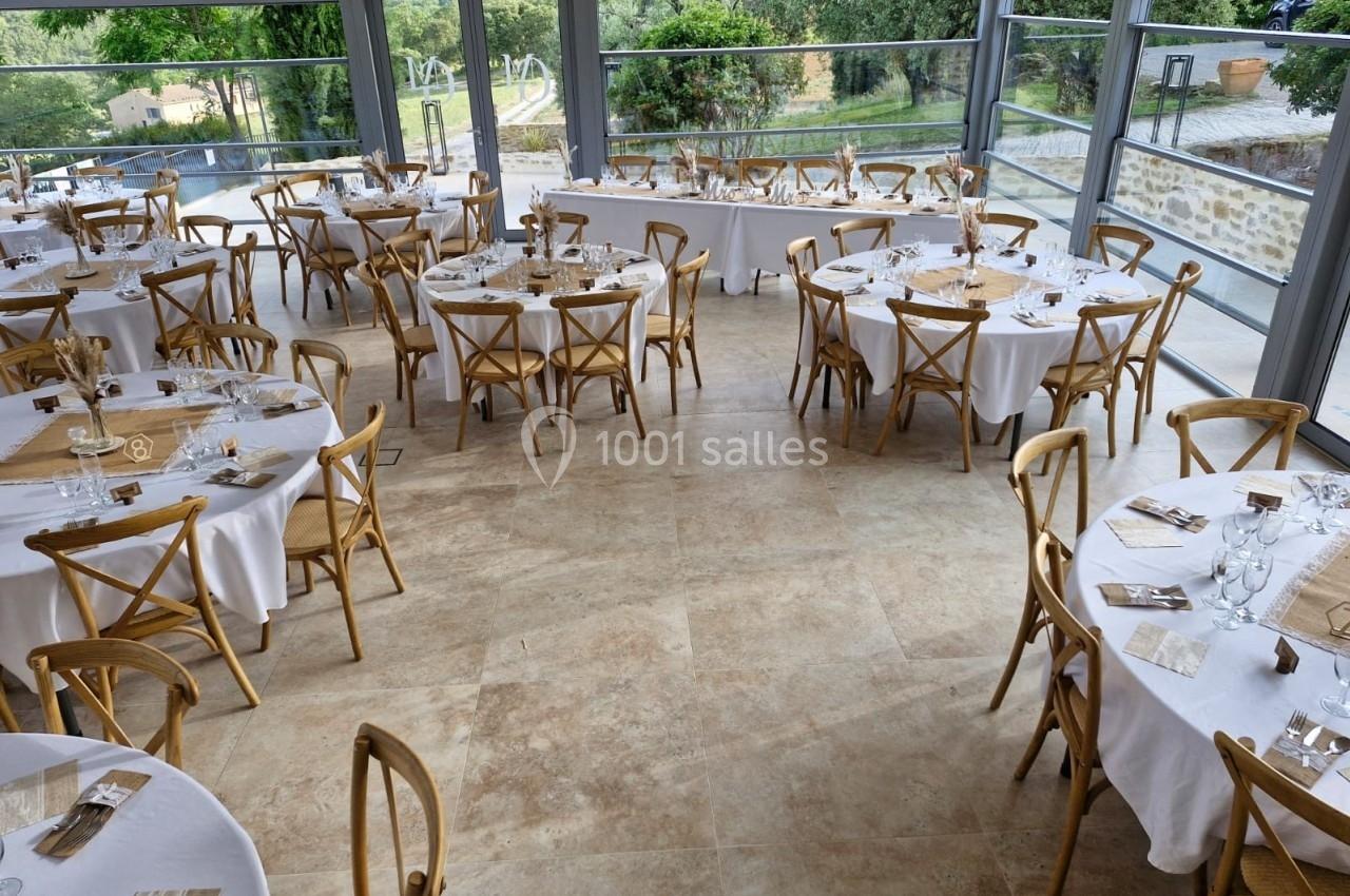 Salle lumineuse avec tables rondes dressées, nappes blanches et chaises en bois, entourée de baies vitrées donnant sur un…