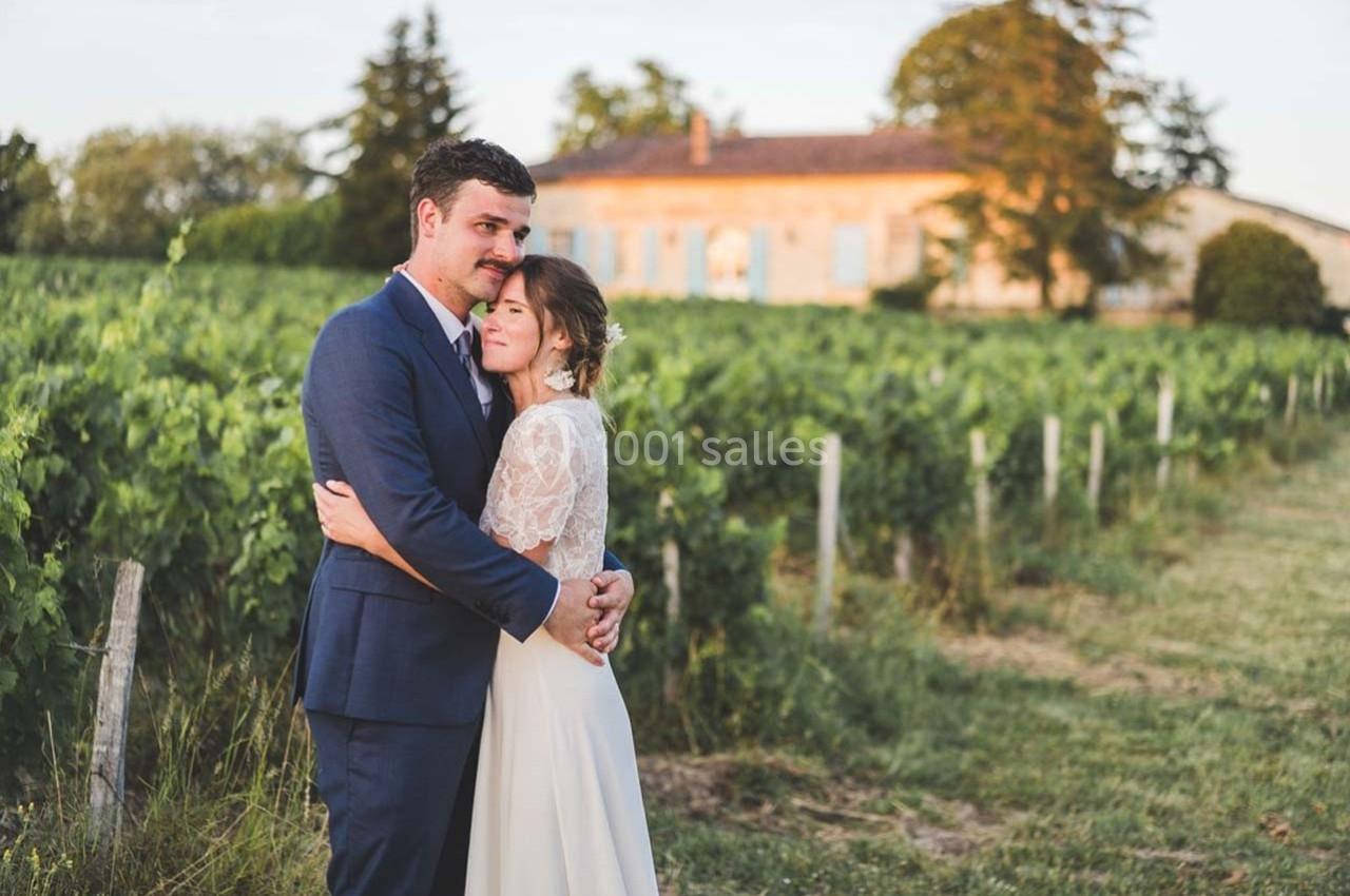 Un couple en tenue de mariage s'enlace devant un vignoble et une maison en pierre au coucher du soleil.