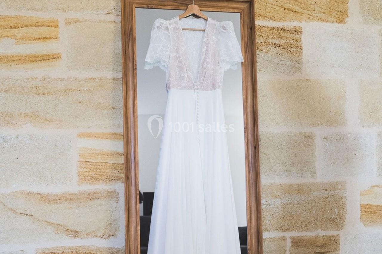 Robe de mariée blanche avec dentelle suspendue devant un miroir encadré en bois, sur un mur en pierre claire.