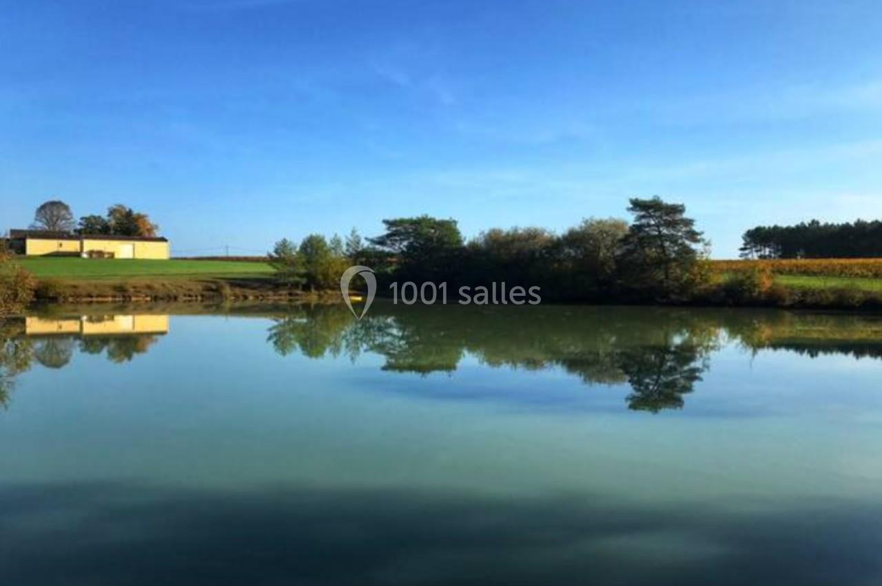 Plan d'eau calme entouré de végétation, avec un bâtiment et des champs sous un ciel bleu dégagé.