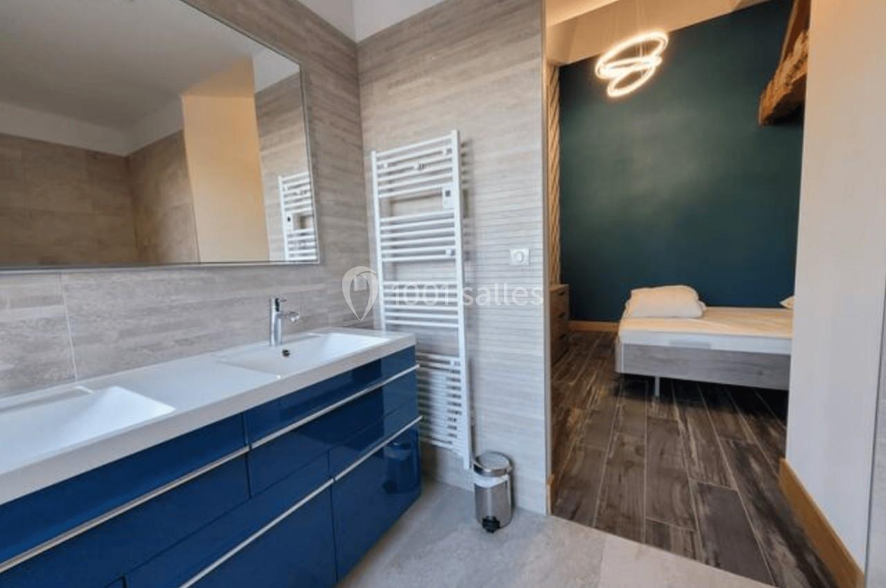 Salle de bain moderne avec meuble bleu, miroir, sèche-serviettes et vue sur une chambre avec lit et mur bleu.