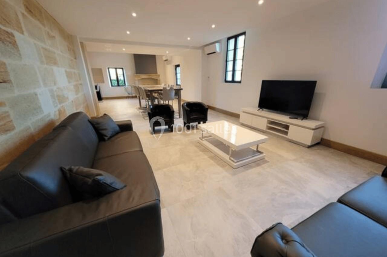 Salon moderne avec canapés en cuir noir, table basse blanche, télévision et espace salle à manger en arrière-plan.