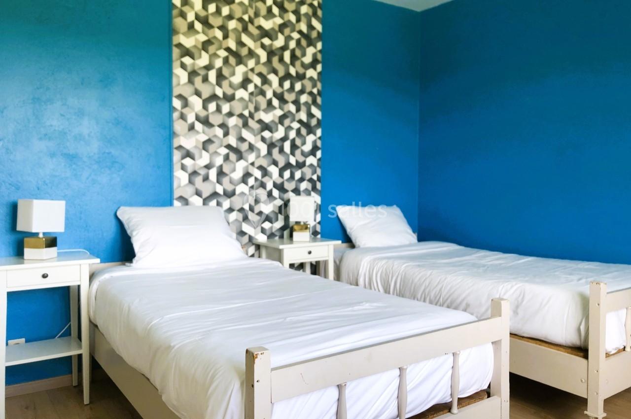 Deux lits simples avec linge blanc, tables de chevet et lampes, dans une chambre aux murs bleus et décor géométrique.