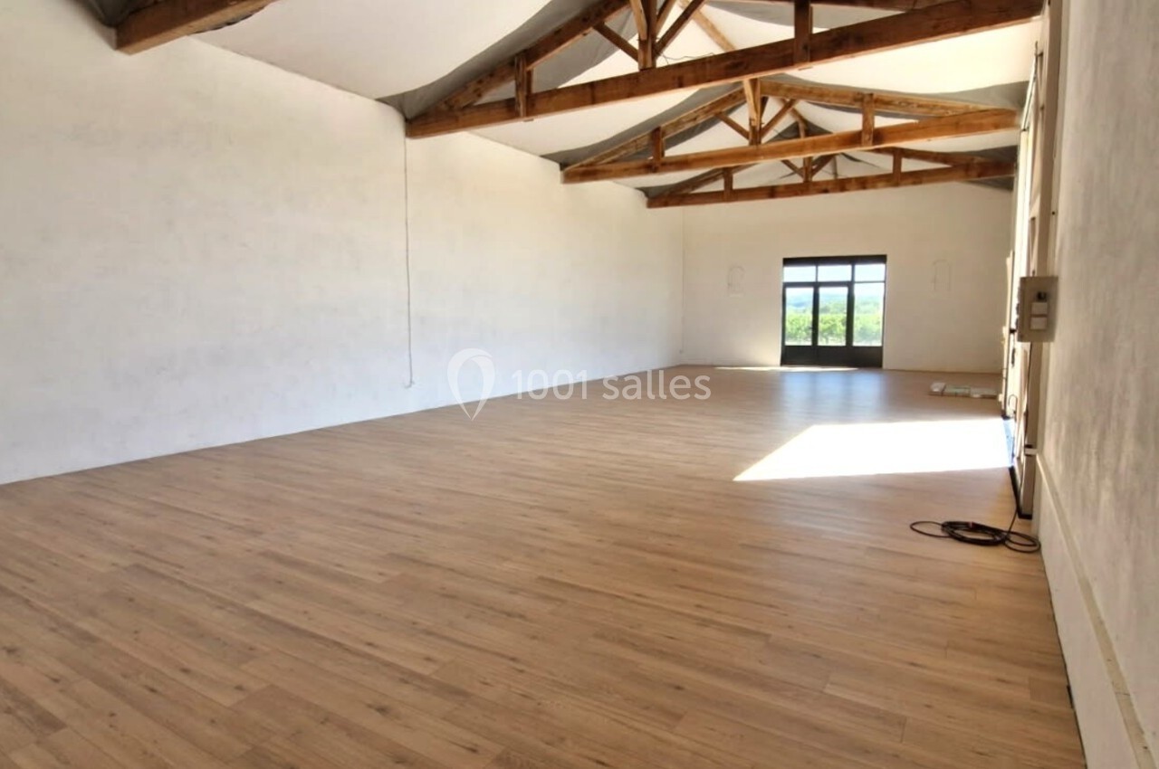 Salle vide avec parquet clair, murs blancs, poutres apparentes au plafond et grande porte vitrée laissant entrer la lumière.