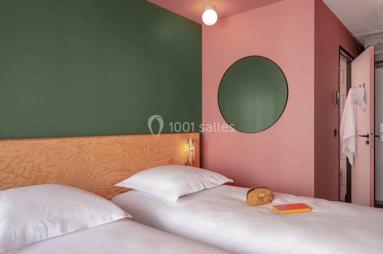 Deux lits simples avec draps blancs dans une chambre aux murs rose et vert, miroir rond et lumière douce.