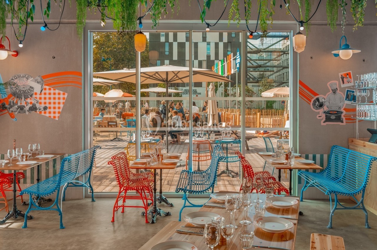 Salle de restaurant colorée avec tables dressées, vue sur une terrasse extérieure avec parasols et mobilier de jardin.