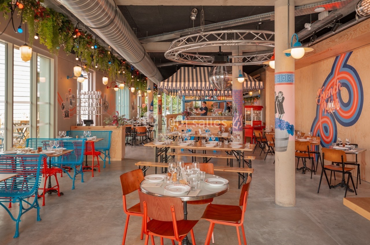Salle de restaurant lumineuse avec tables dressées, décor coloré, plantes suspendues et éléments industriels.