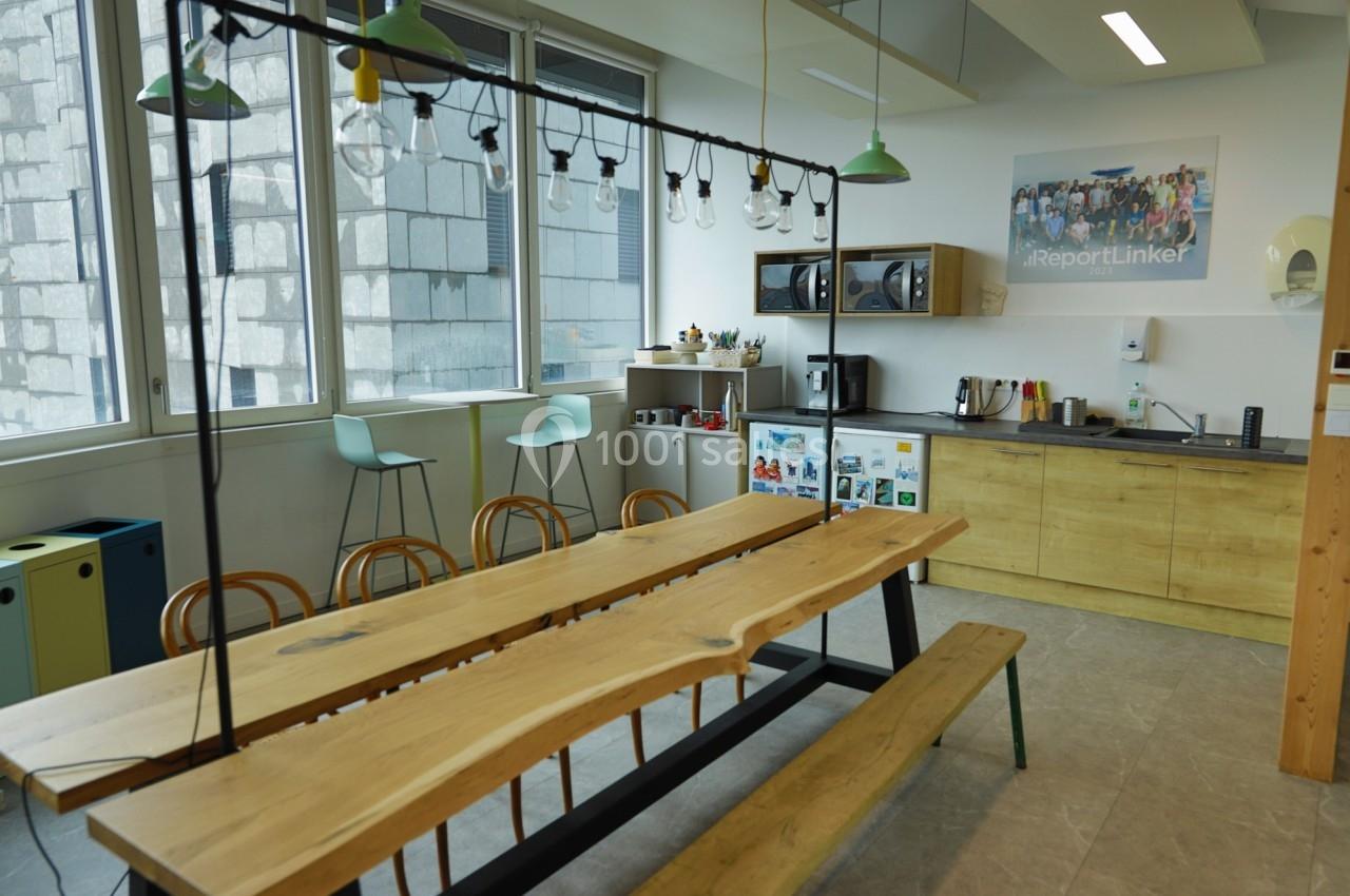 Salle de pause lumineuse avec table en bois, chaises, cuisine équipée et grandes fenêtres donnant sur un mur extérieur.