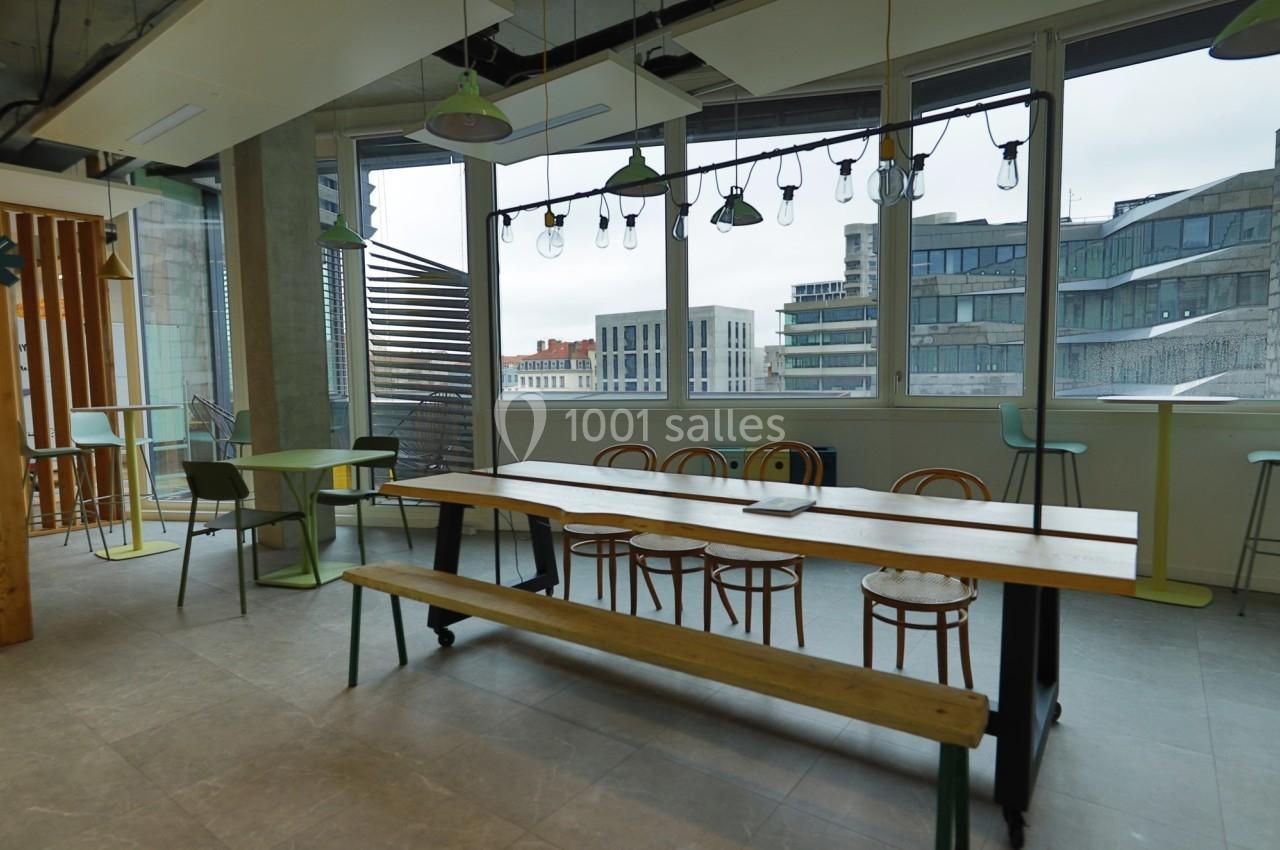 Espace de pause lumineux avec grande table en bois, chaises, tabourets et vue sur des immeubles modernes.