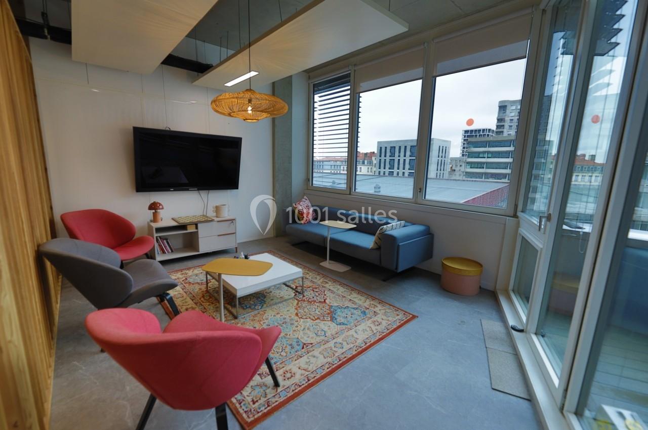 Espace de détente lumineux avec fauteuils colorés, table basse, tapis et grande fenêtre donnant sur des immeubles.