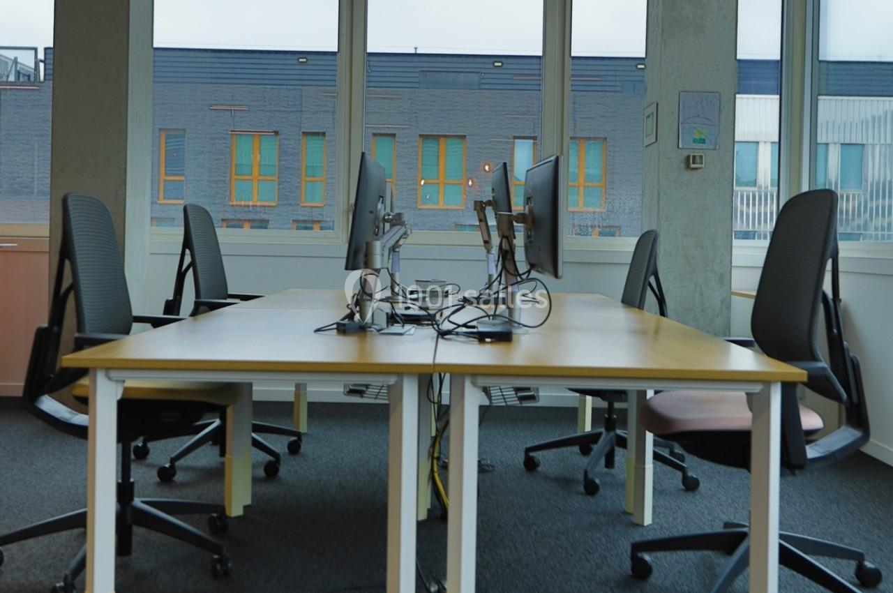 Quatre bureaux équipés d'écrans et de chaises dans un espace de travail lumineux avec de grandes fenêtres.