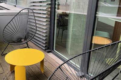 Deux fauteuils noirs et une table jaune sur une terrasse en bois, près de baies vitrées et de stores.