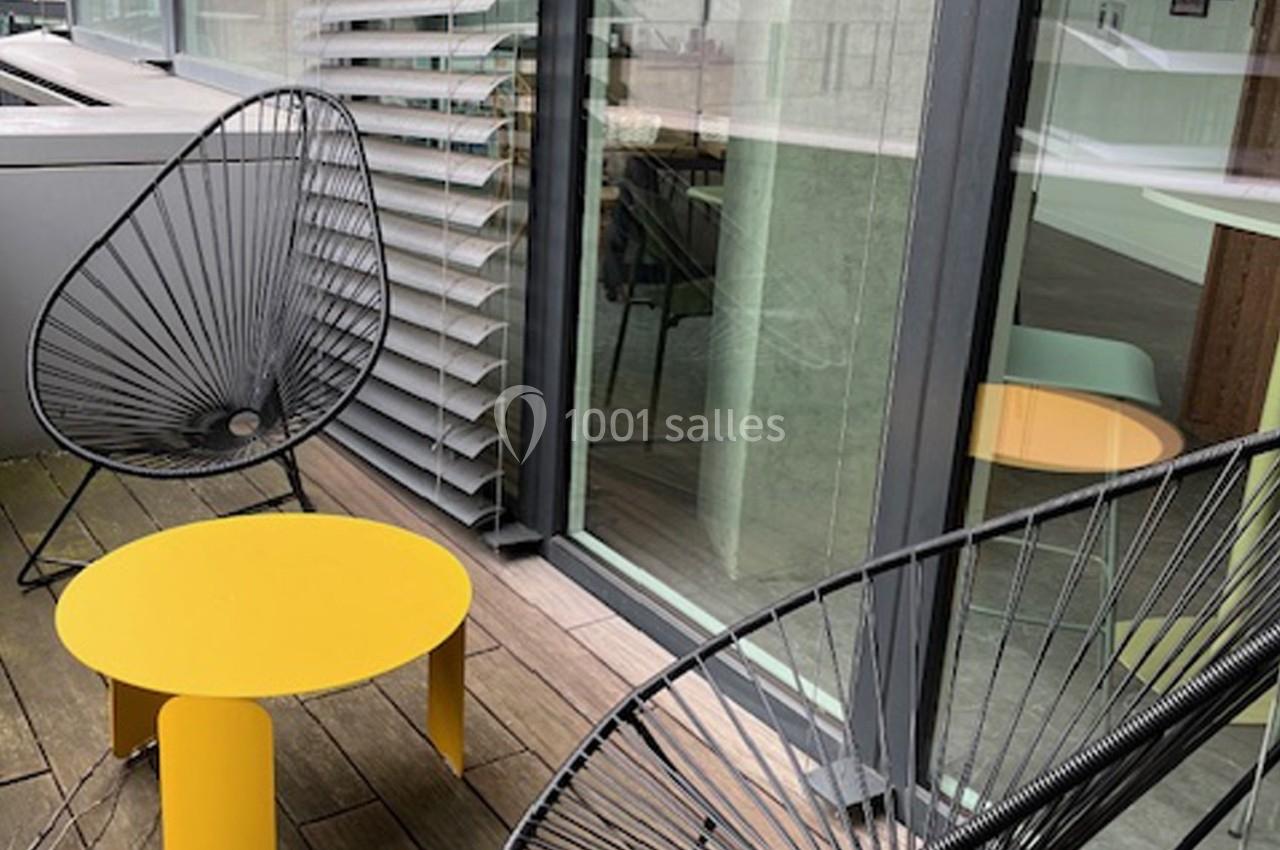 Deux fauteuils noirs et une table jaune sur une terrasse en bois, près de baies vitrées et de stores.