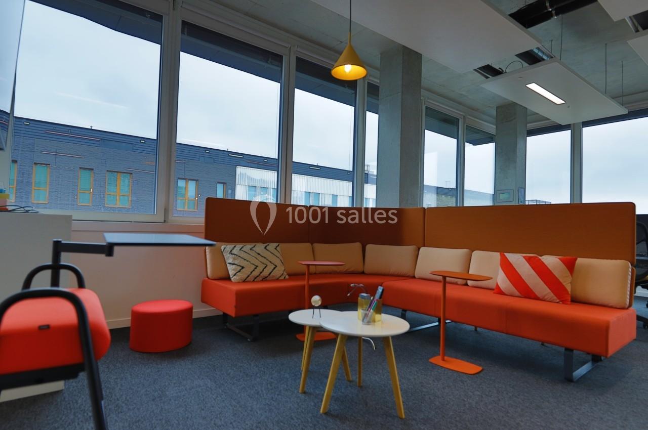 Espace de détente avec canapé orange, table basse blanche, pouf rouge et grandes fenêtres donnant sur l'extérieur.