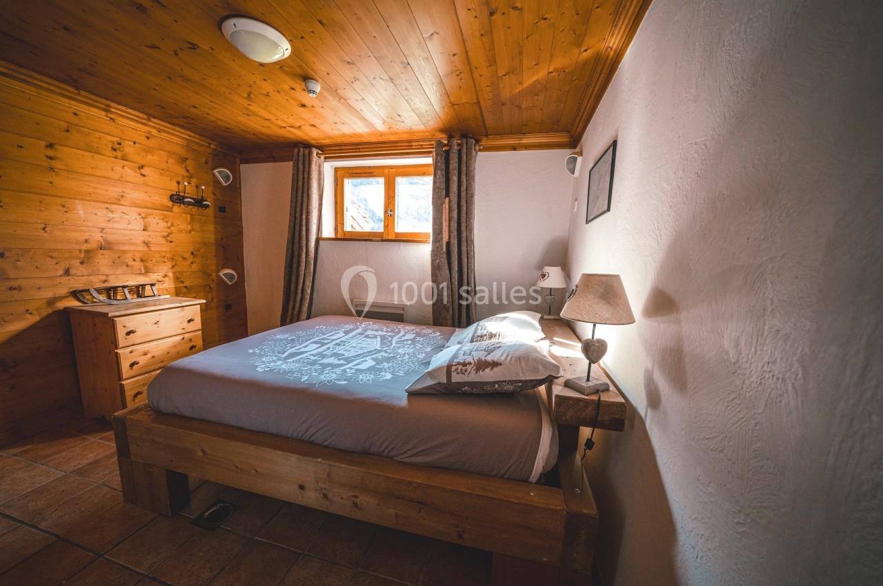 Chambre en bois avec un lit double, une commode, une lampe de chevet et une fenêtre laissant entrer la lumière naturelle.