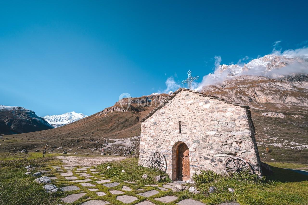 Petite chapelle en pierre avec une croix sur le toit, entourée de montagnes sous un ciel bleu dégagé.