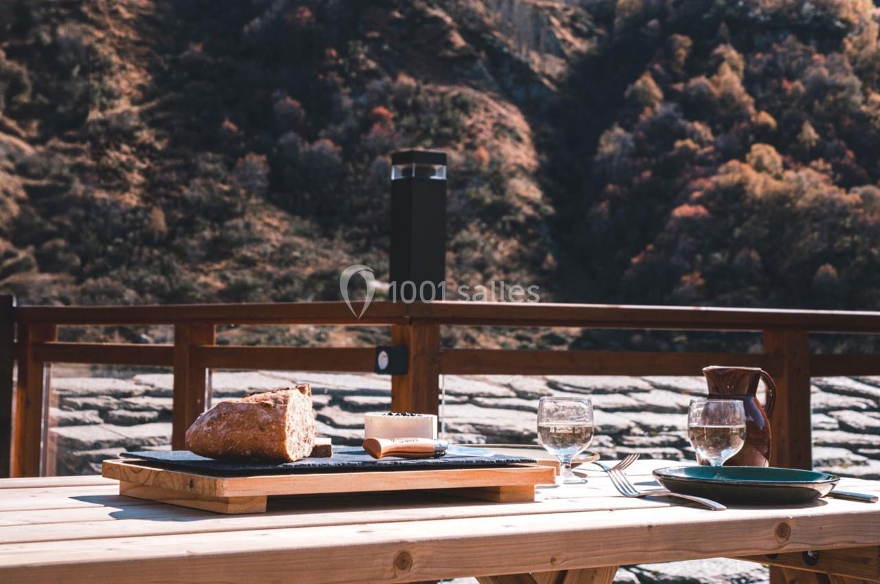 Table en bois avec pain, fromage, verres et couverts, située en extérieur avec vue sur un paysage montagneux automnal.