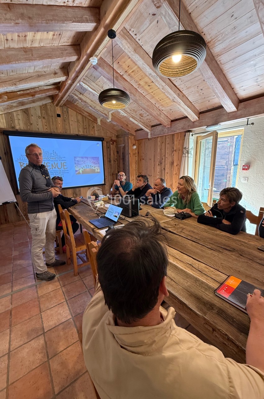 Un groupe de personnes assises autour d'une table en bois écoute un présentateur dans une salle au style rustique.