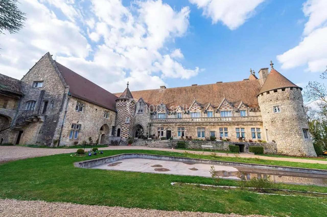 Château en pierre avec tours et toits en tuiles, entouré d'une pelouse et d'une allée gravillonnée, sous un ciel…