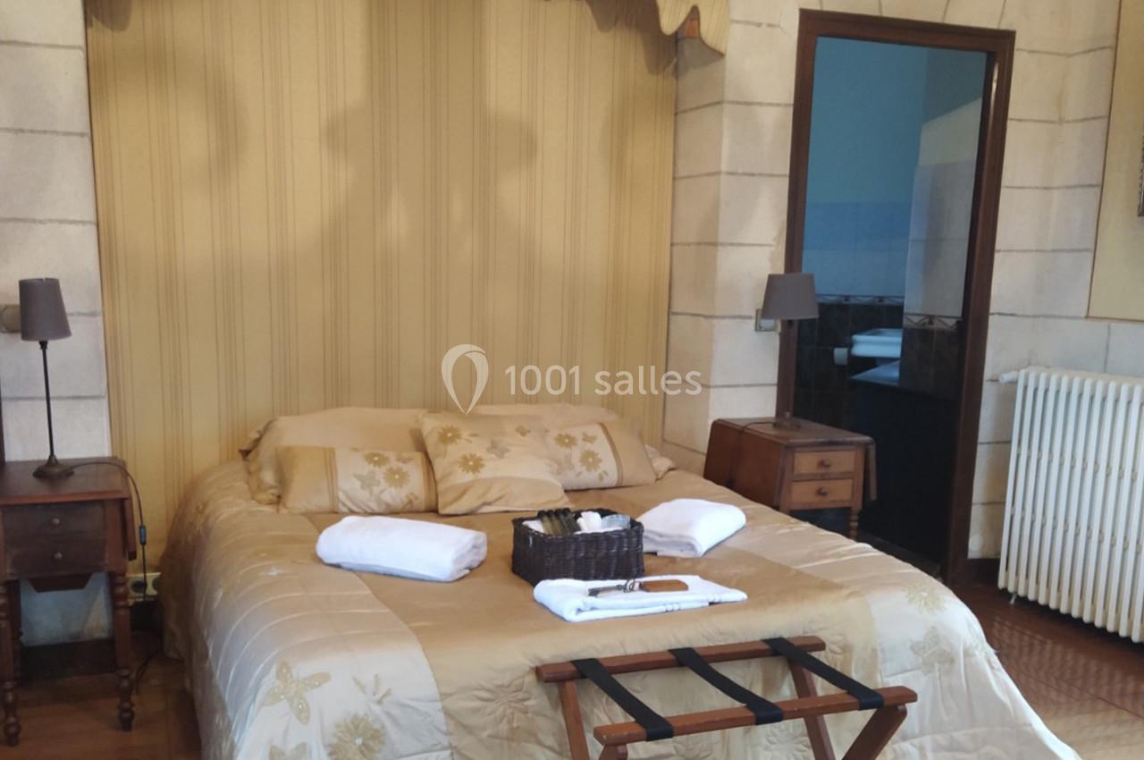 Lit double avec couvre-lit beige, deux tables de chevet, une corbeille d'accueil et une salle de bain visible en arrière…