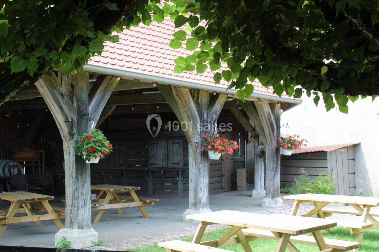 Abri en bois avec toit en tuiles, tables de pique-nique et jardinières suspendues, entouré de verdure.