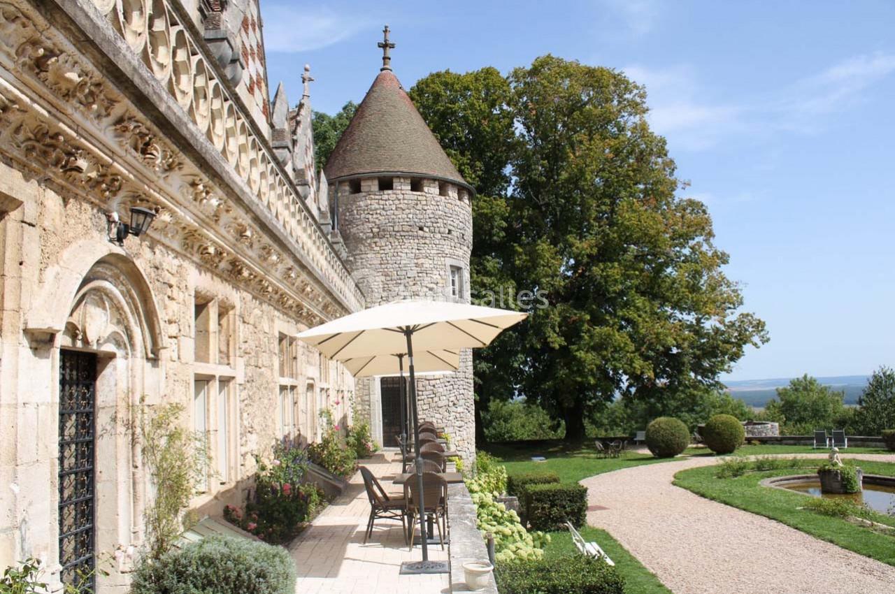 Façade en pierre d'un château avec une tour ronde, terrasse aménagée et jardin verdoyant sous un ciel dégagé.