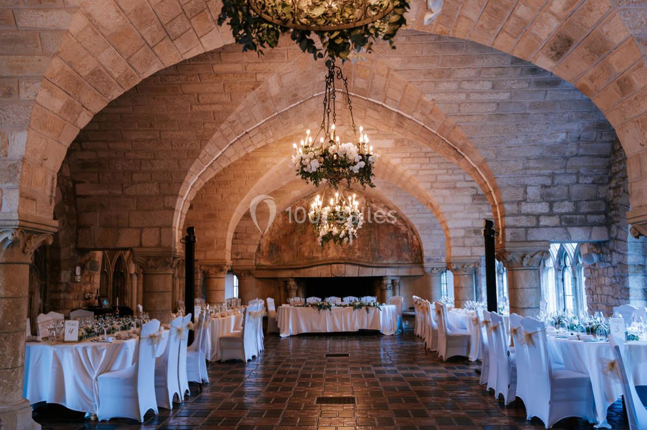 Salle voûtée en pierre avec tables dressées pour un événement, éclairée par des lustres et la lumière naturelle.