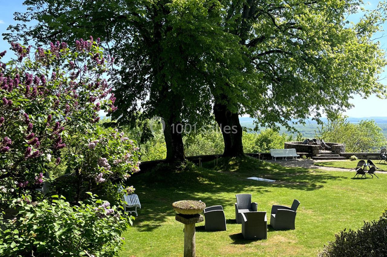 Jardin verdoyant avec chaises en rotin, grande table en pierre, lilas en fleurs et vue dégagée sur l'horizon.