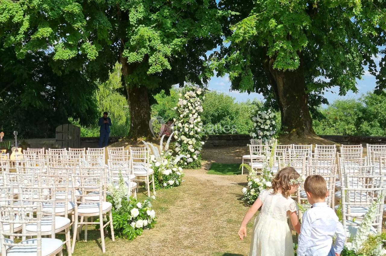 Allée centrale décorée de fleurs blanches entre des rangées de chaises, menant à une arche florale sous des arbres.