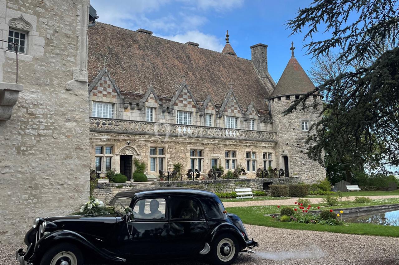 Voiture ancienne noire garée devant un château en pierre avec toit en tuiles et tour, entouré de jardins.