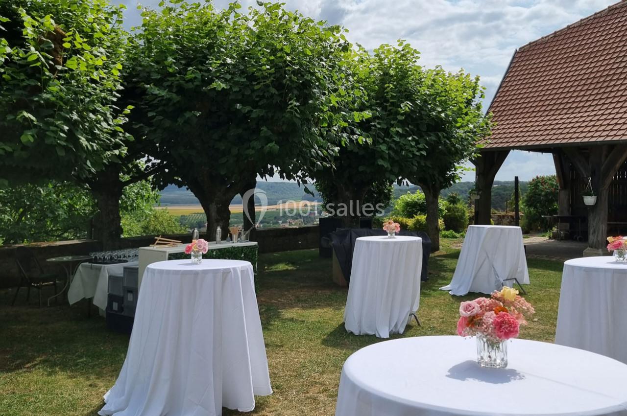 Tables hautes recouvertes de nappes blanches et décorées de fleurs, disposées dans un jardin ombragé.
