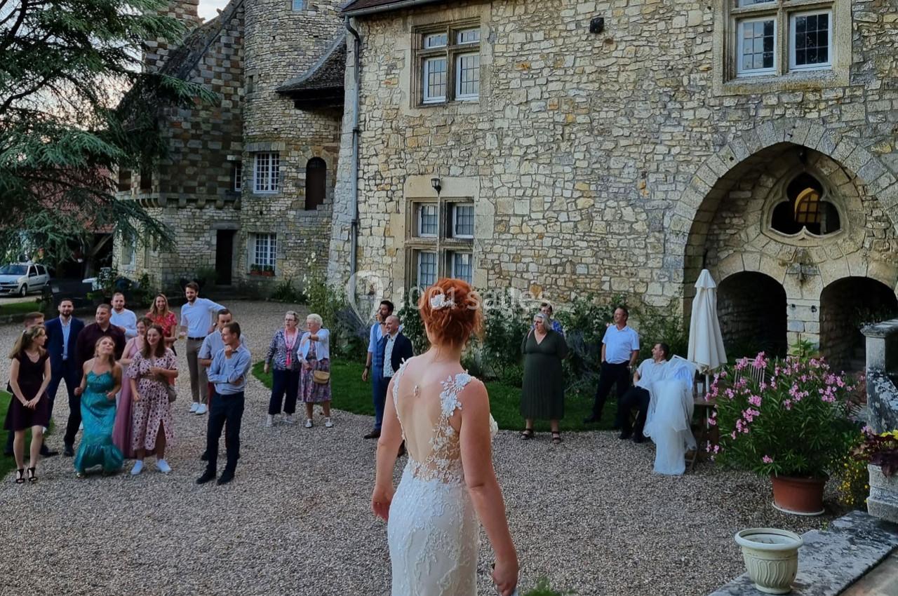 Une mariée en robe blanche fait face à des invités rassemblés dans la cour d'un bâtiment en pierre.