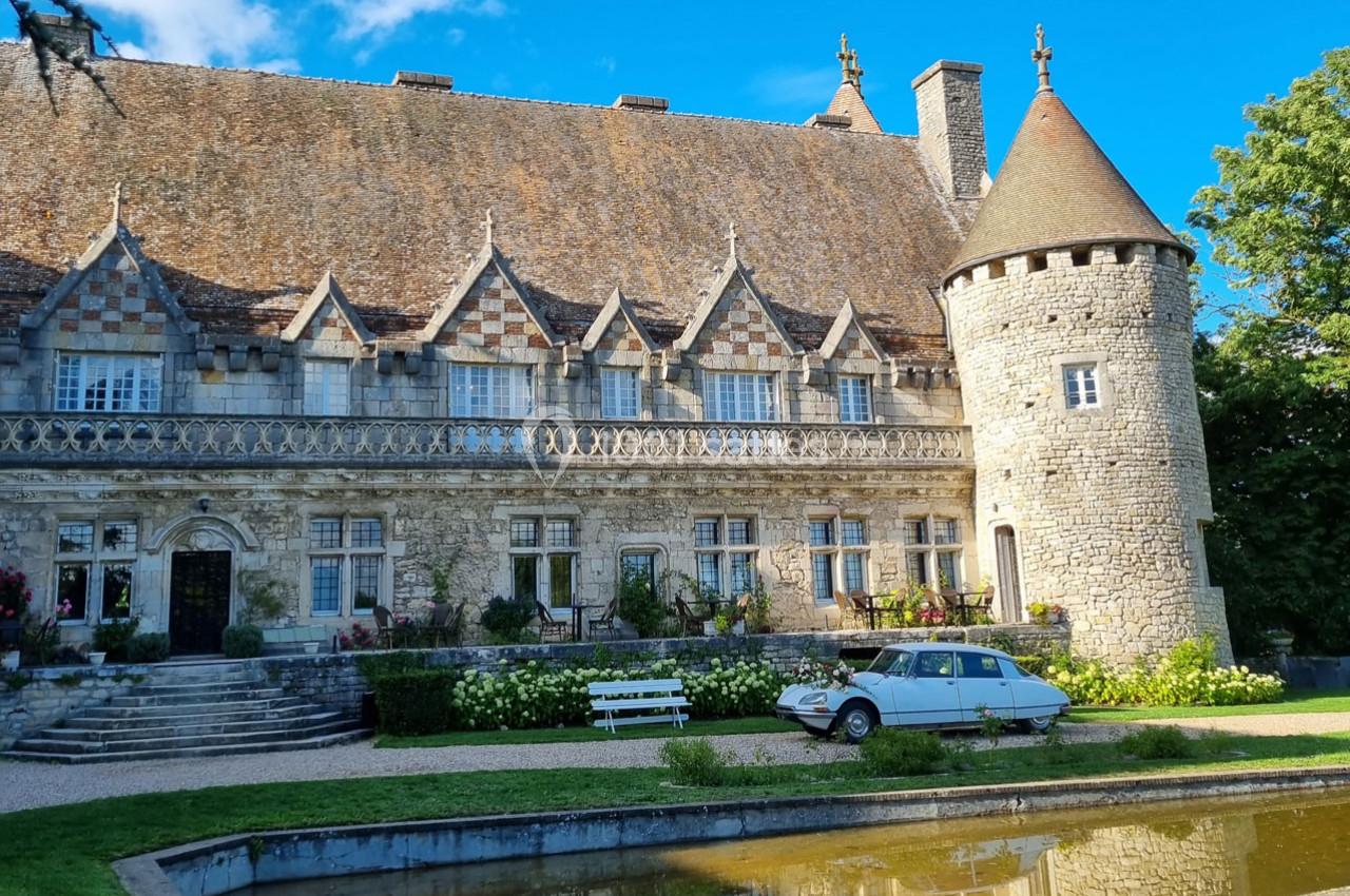 Façade d'un manoir en pierre avec une tour ronde, une voiture ancienne et un bassin au premier plan.