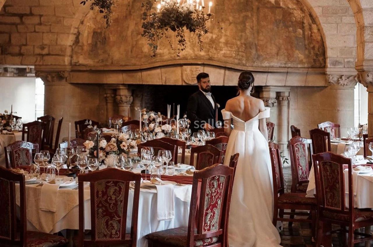 Une mariée en robe blanche et un homme en costume dans une salle de réception élégante avec tables dressées.