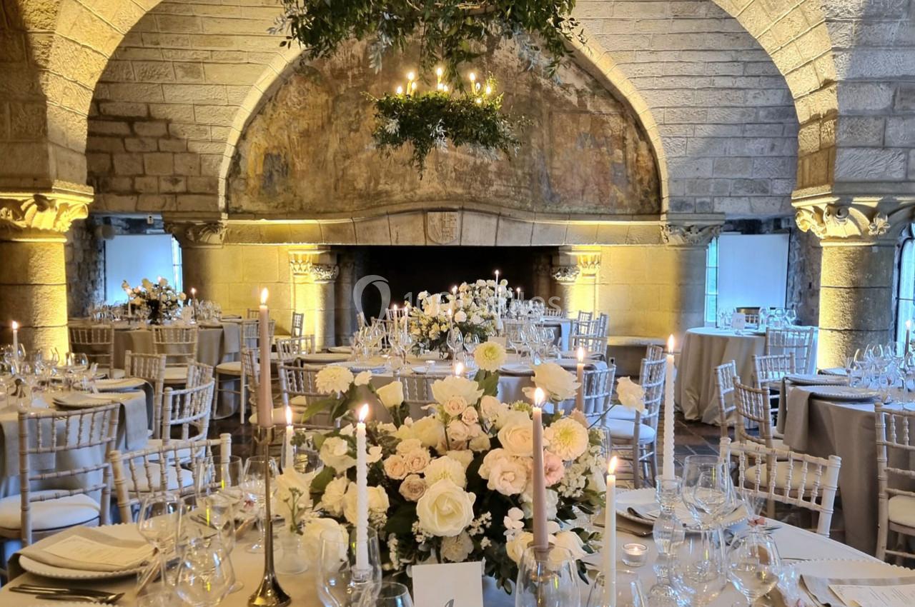 Salle de réception élégante avec tables décorées de fleurs blanches et bougies, sous des voûtes en pierre.