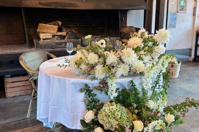 Salle en bois avec de grandes fenêtres, tables en bois décorées pour un repas, chaises pliantes et éclairage suspendu.