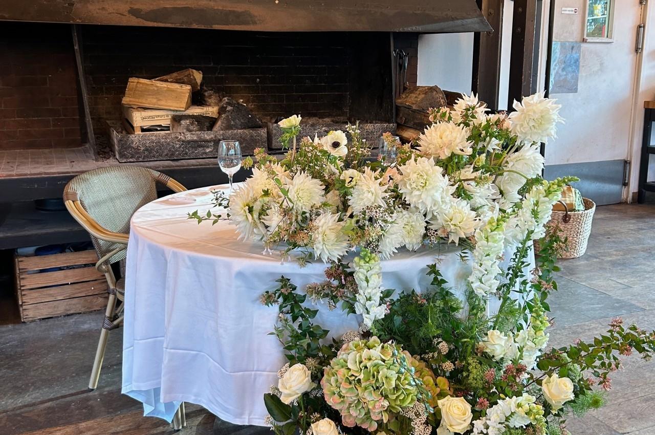 Table décorée de fleurs blanches et vertes devant une cheminée en pierre dans une salle rustique.