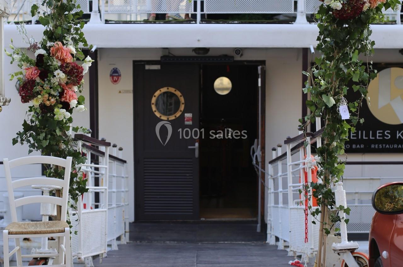 Entrée d'un bateau décorée d'une arche florale, avec des personnes discutant sur le pont supérieur.