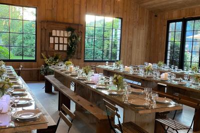Salle en bois avec de grandes fenêtres, tables en bois décorées pour un repas, chaises pliantes et éclairage suspendu.