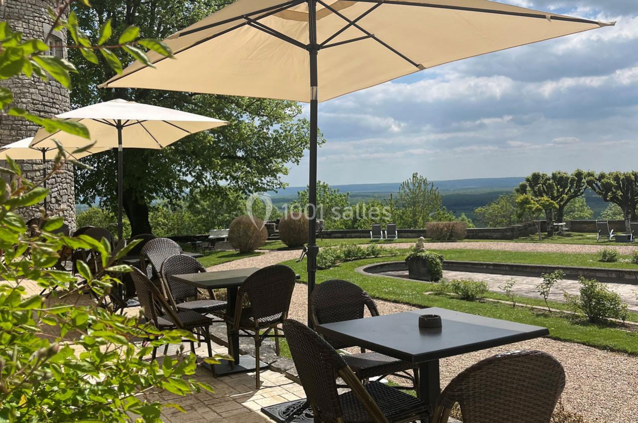Terrasse avec tables et chaises en osier sous des parasols, offrant une vue dégagée sur un paysage verdoyant.