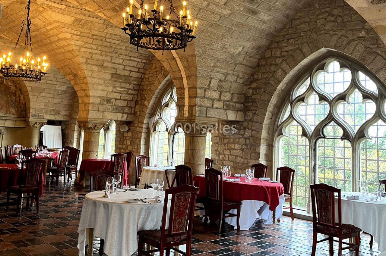 Salle de restaurant élégante avec des tables dressées, des chandeliers suspendus et de grandes fenêtres en arc gothique.