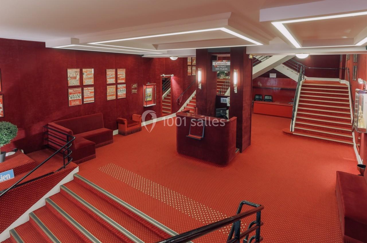 Hall d'entrée d'un bâtiment avec moquette rouge, comptoir d'accueil, escaliers et affiches encadrées sur les murs.