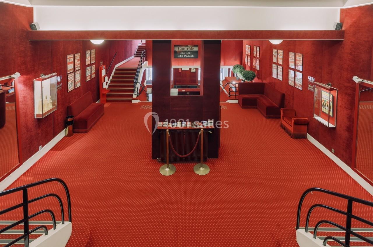 Hall d'entrée d'un théâtre avec moquette rouge, affiches encadrées sur les murs et comptoir central avec brochures.