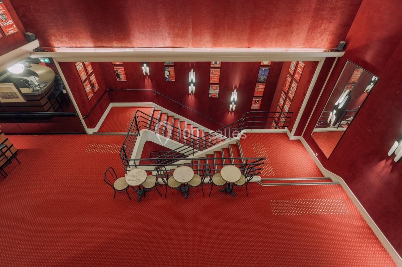 Escalier central entouré de murs rouges, avec tables rondes et chaises noires dans un espace intérieur lumineux.