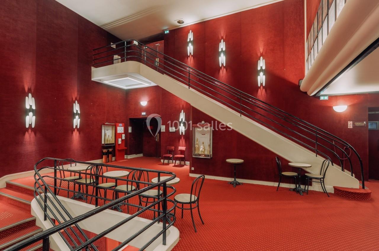 Escalier central dans un hall intérieur aux murs et moquette rouges, avec tables, chaises et éclairage mural.
