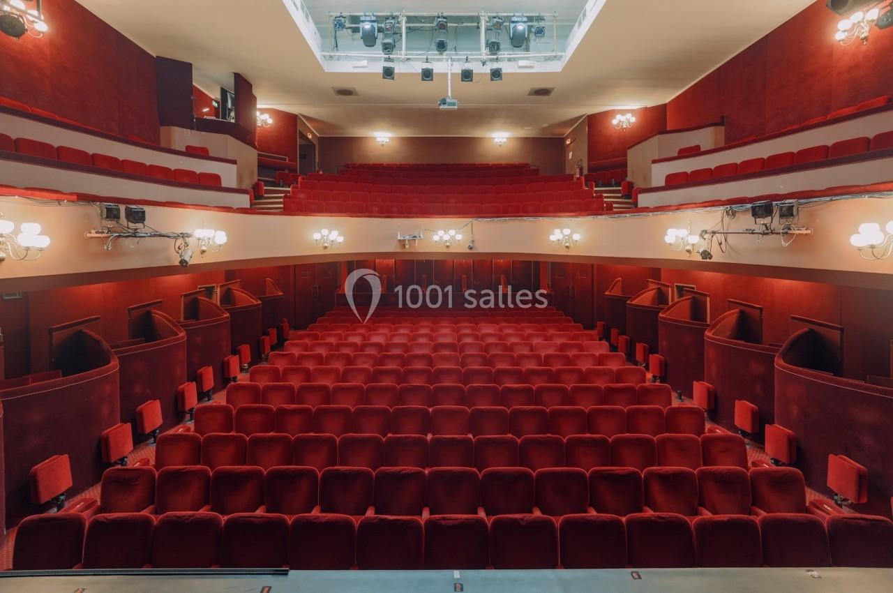 Salle de théâtre vide avec des rangées de sièges rouges, éclairée par des lumières au plafond et sur les côtés.