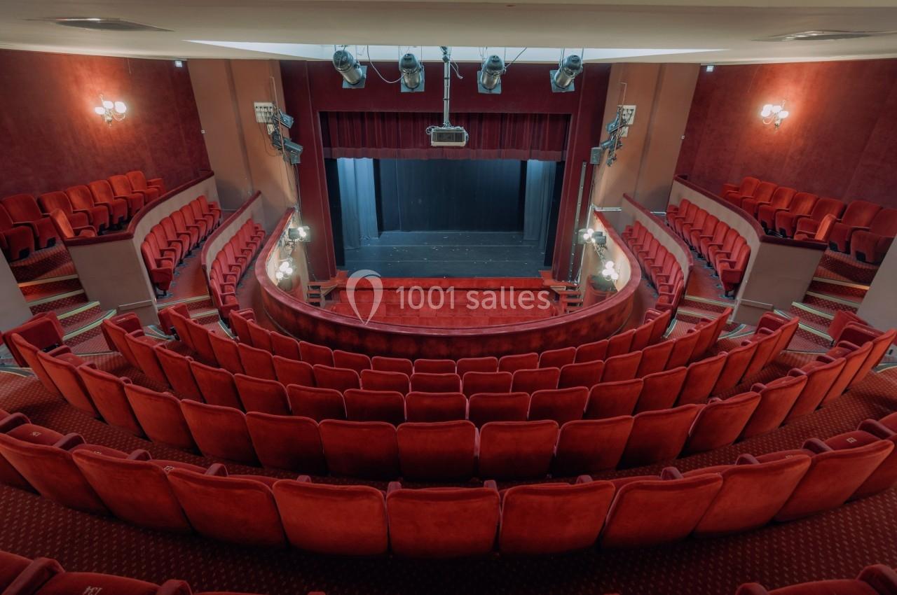 Salle de théâtre vide avec des rangées de fauteuils rouges, une scène et des projecteurs suspendus au plafond.