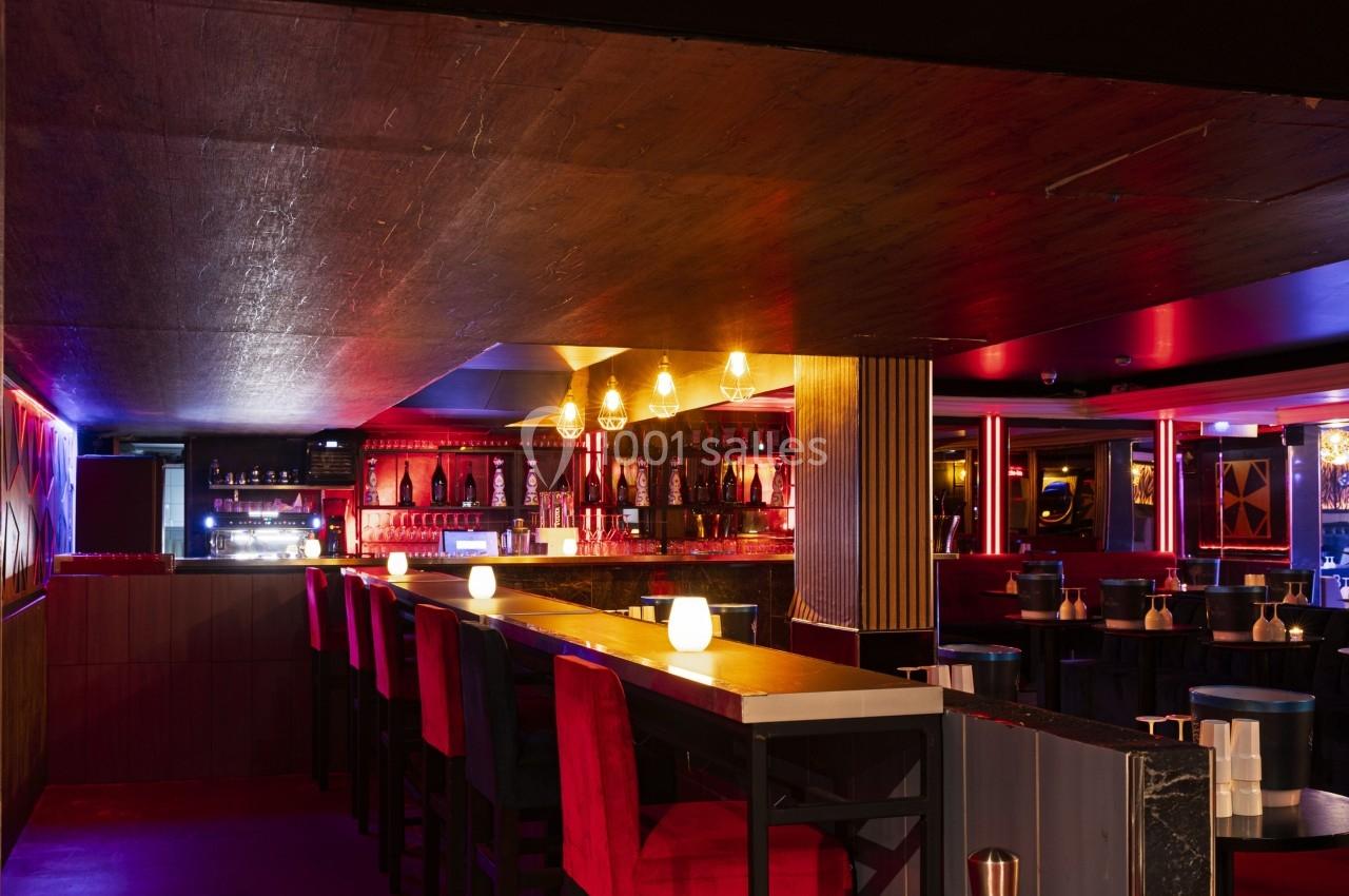 Intérieur d'un bar avec un comptoir éclairé, des tabourets rouges et une ambiance lumineuse tamisée.