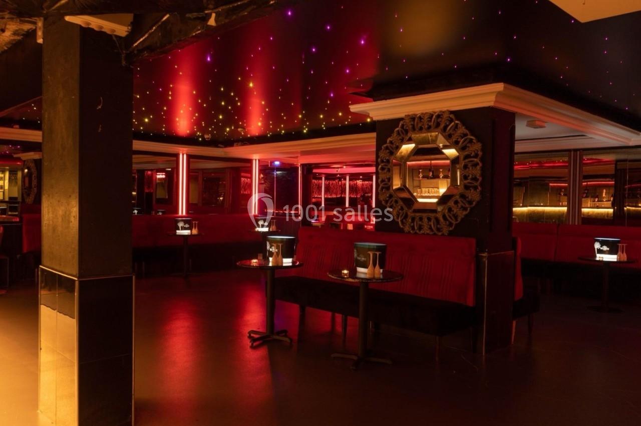 Salle de bar ou club avec éclairage tamisé rouge, banquettes, tables basses et miroir décoratif sur un mur.